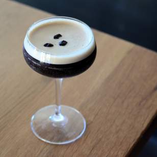 Close-up of 101 Craft Kitchen espresso martini with three coffee beans on top, served in a coupe glass on a wooden table.