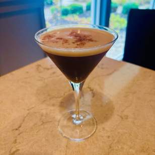 Pumpkin Spice Espresso martini with a dusting of cocoa on a marble table at Gallo's in Dublin, Ohio, with a window view in the background.