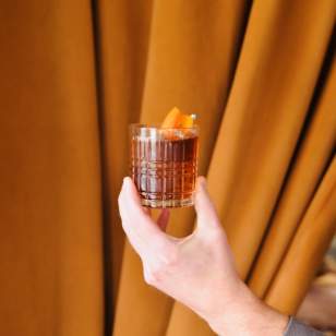 A hand holding a classic whiskey cocktail in a textured glass, garnished with an orange peel, against a backdrop of warm, golden-brown curtains.