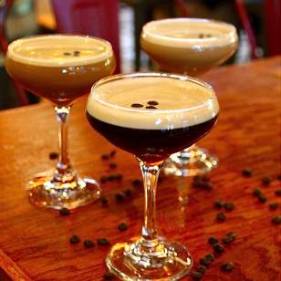 Three espresso martinis at Market Bar on a wood table with coffee beans, red chairs blurred in the background.