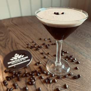 Salted Cold Brew Espresso martini garnished with three coffee beans on a wooden table surrounded by scattered beans and a North High Brewing coaster.