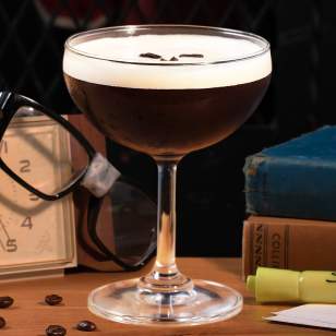Close-up of PINS Mechanical "All Nighter" espresso martini with coffee beans on top, surrounded by books, glasses, and coffee beans on a wood table.