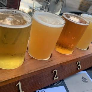 Beer Flight (North High Brewing): A wooden tray labeled with numbers 1 to 4 holds four small glasses of beer, each showcasing a different hue, from pale gold to rich amber. The beers appear freshly poured, with bubbles rising to the surface. A relaxed outdoor setting with a faint reflection of sunlight and patio furniture in the background enhances the casual atmosphere.