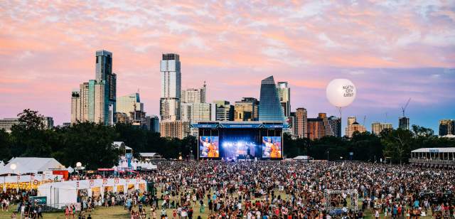 Aerial shot of Zilker Park during ACL festival with a sea of fans crowding the main stage, a cotton candy colored sky with the Austin skyline as the backdrop.