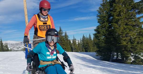 A skier uses adaptive ski equipment with a companion to ski the slopes. Snow, trees and blue sky in the backdrop.