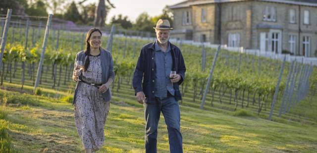 A couple walk through the vines, with Leonardslee House and a sculpture in the background