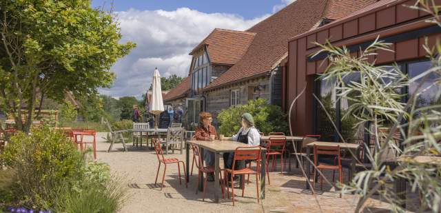 A couple sit at a table in a sunny courtyard surrounded by plants, trees and the Knepp kitchen buildings