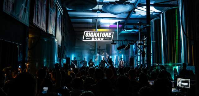 A lively concert scene in a dimly lit brewery, with a crowd facing a band under a neon "Signature Brew" sign, brewing vats are on either side of the audience. The atmosphere is energetic and vibrant.