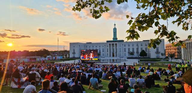 Fellowship Square lawns and gardens with crowds watching a large screen showing a sporting event.Sunset backdrop, tree branches in the foreground and Waltham Forest Town hall and clock tower in the back ground.