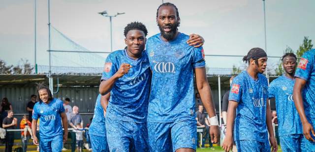 Two football players in blue William Morris print kits, smiling and arm-in-arm, walk towards the camera on a sunny day. Other players and spectators are in the background.