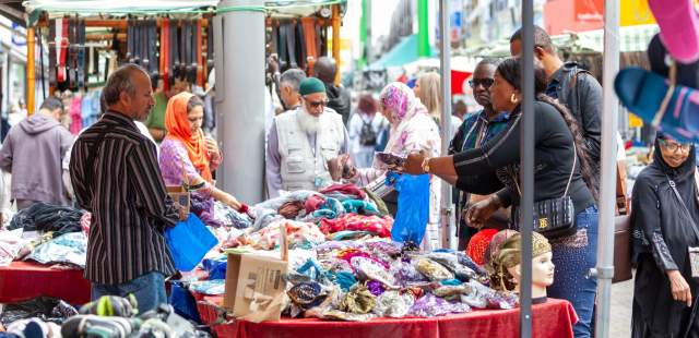 A bustling outdoor market scene with diverse shoppers examining colourful fabrics on a table. Mannequin heads display headscarves, creating a lively, multicultural atmosphere.