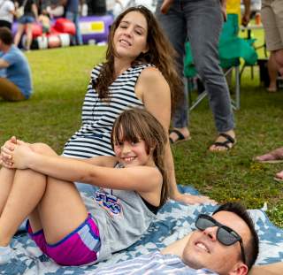A family relaxes on a picnic blanket at Fayetteville, NC's Greek Festival, with a smiling young girl in the center and inflatable bounce houses in the background.