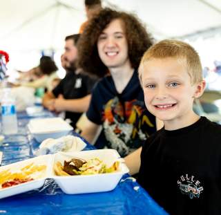 A young boy smiles at the camera while sitting at a long blue-covered table, with a tray of fries and gyro meat in front of him and festivalgoers enjoying food in the background.