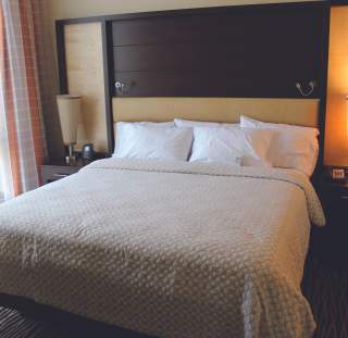 Hotel guest room with a king bed, neutral bedding, bedside tables with lamps, and natural light from a window.