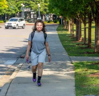 A young woman smiles confidently while walking alone on a tree-lined sidewalk in downtown Fayetteville, NC—backpack on, keys in hand, ready to explore.