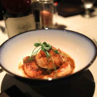 A white bowl with blue trim contains steamed scallops with seasoning and a leaf of garnish