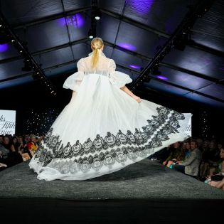 A model wearing a long and billowy white dress walks down the runway lined with people on both sides, during Fashion Week El Paseo 2023.