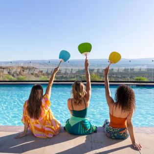 Three girl friends holding up fans and posing for a photo poolside at The Ritz-Carlton, Rancho Mirage.