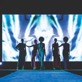 The silhouette of five women standing on the runway in front of a large screen.