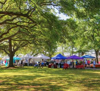 Visitors explore a vibrant outdoor market in Sunset Beach, NC, with colorful vendor tents under shady trees on a bright, sunny day.