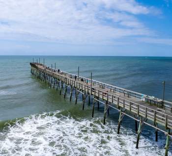 Long wooden fishing pier extending over the Atlantic Ocean in North Carolina’s Brunswick Islands, with people walking along the pier and gentle waves breaking below under a clear blue sky.
