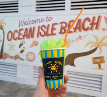 a cup of green italian ice from Sunset Slush pictured in front of a mural on Ocean Isle Beach