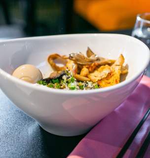 A bowl of ramen on a dark table, featuring a soft-boiled egg, mushrooms, and green onions. Nearby, there is a glass of water and black chopsticks on a pink napkin.