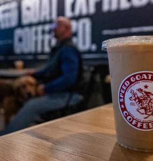 A clear plastic cup with iced coffee sits on a wooden table in a cafe. The logo reads "Speed Goat Espresso." A blurred background shows a person and a dog.