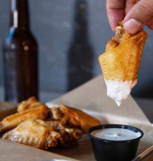 Person dipping wing into ranch with a basket full of wings and a beer in the background.