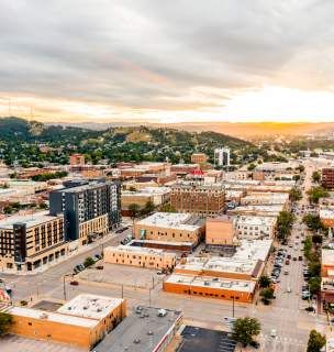 Aerial view of downtown Rapid City at sunset, with downtown streets lined by low-rise buildings, cars, and parking lots. Rolling green hills surround the city, while dramatic clouds stretch across the sky and golden sunlight glows on the horizon.