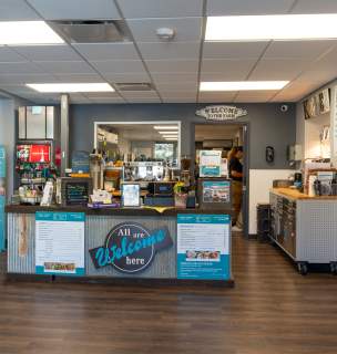 Cafe interior with a welcoming atmosphere. The counter has a sign reading "All are Welcome here." Menu boards and decor add warmth to the cozy space.