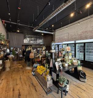 Spacious store interior with wooden floors and brick walls. Shelves are stocked with various goods, including plants and signs. Refrigerators line the right wall. Warm, inviting ambiance.