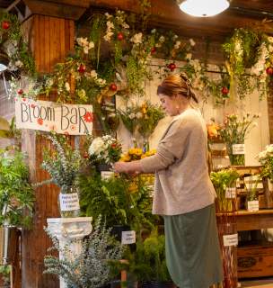 Woman arranging flowers at Victoria's Garden "Bloom Bar." Lush greenery, colorful blooms, rustic decor and a warm, inviting atmosphere.