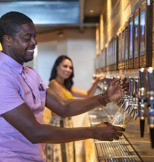 Male pouring his own beer from a wall tap with female in the background doing the same.