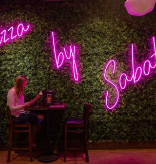 Woman sitting a bar height table reading a restaurant menu. A neon pink restaurant sign illuminates the background.