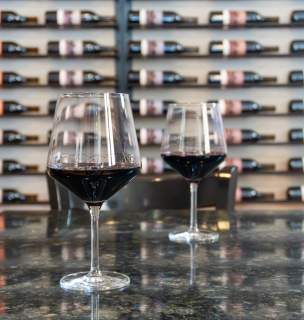 Two glasses of red wine in wine glasses sit on a countertop. A wall of colorfully-labeled wine bottles lay horizontally on a wall rack in the background, slightly out of focus.