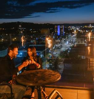 Couple enjoying drinks at a rooftop bar beside a glass fire feature, overlooking the glowing city lights of Rapid City at dusk. The pair sits at a small table, toasting mugs while the skyline and Black Hills foothills stretch into the evening background.