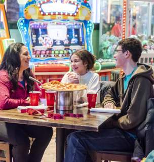 Family of three sitting around a table eating pizza with arcade games in the background at Press Start in Rapid City, SD