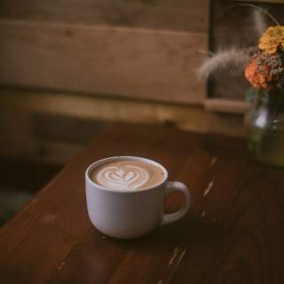 A latte served in a white cup sits on a table next to a vase with orange and yellow flowers.