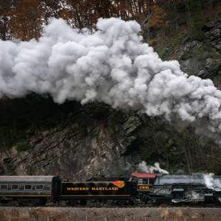 The Western Maryland Scenic Railroad steam engine billows smoke from the engines and pulls along a passenger car near a mountain wall.