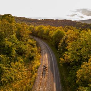 Four passengers peddle along a rail line on peddle bikes with the trees, sky and clouds in the background at sunset.