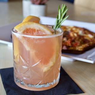 A refreshing cocktail garnished with lemon and rosemary sits on a black napkin, with a colorful dish in the background.