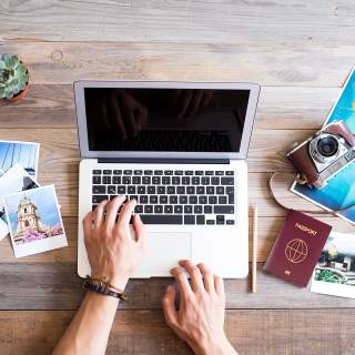 Travel documents and photos sit on a table as a person uses a laptop.