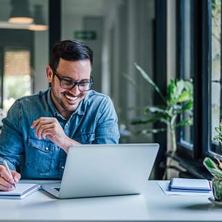 A man takes notes on a notepad while smiling at his laptop screen.