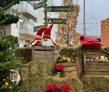 En juledekorert scene i sentrum av Lyngdal, med halmballer, trekasser fylt med julepynt, to nisser, røde julestjerner og et grønt veiviserskilt som peker til lokale attraksjoner. Små lyslenker og et tre i bakgrunnen gir førjulsstemning.