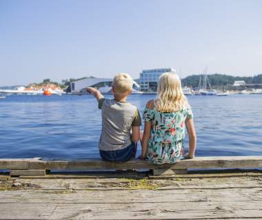 Two children sitting on a dock, pointing towards a building