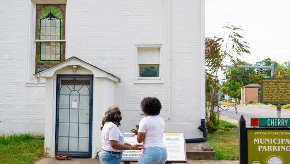 Two women are standing and holding a phone in front of the Kentucky Black Trailblazers plaque for The First Black Baptist Church located in downtown Elizabethtown.
