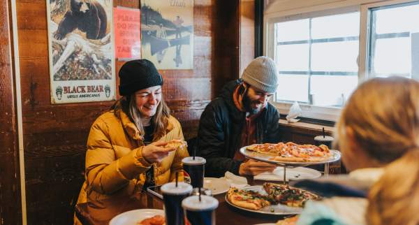Friends in winter hats and jackets eat pizza and drink sodas at a cozy, wood-paneled restaurant