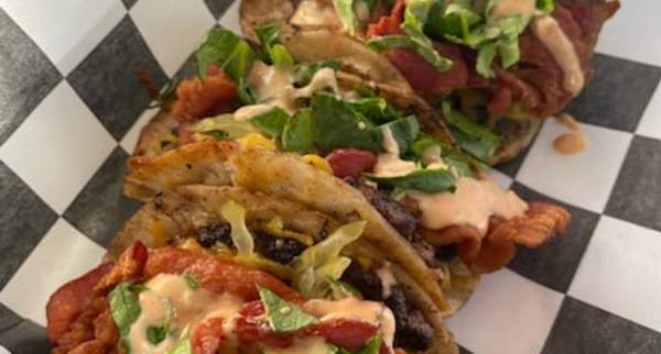 Street tacos topped with grilled meat, bacon, fresh greens, and creamy sauce served in a paper-lined basket
