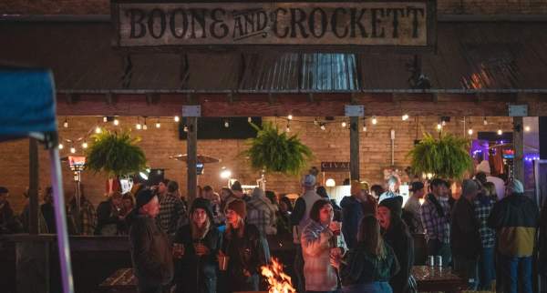 A crowd gathers at Boone & Crockett for an outdoor event at night. People in flannel and jackets socialize with drinks around a fire pit under string lights and hanging plants. The rustic bar sign hangs above the scene, creating a cozy festival atmosphere.